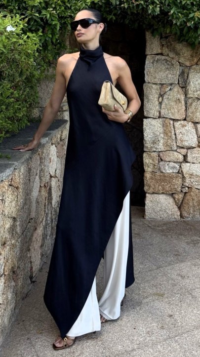 Mulher em pé usando um vestido longo preto com calças brancas, segurando uma bolsa dourada e usando óculos escuros.