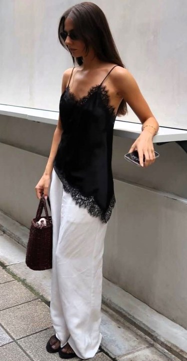 Mulher em pé usando uma blusa preta de seda com rendas e calças brancas largas, segurando uma bolsa marron e um celular.