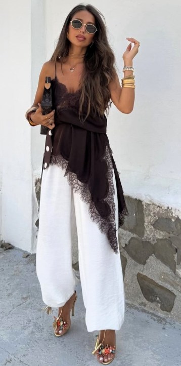 Mulher com cabelo longo e ondulado, vestindo uma blusa preta com detalhes em renda e calças brancas largas. Ela usa óculos de sol e tem uma pose confiante, segurando uma bolsa preta.