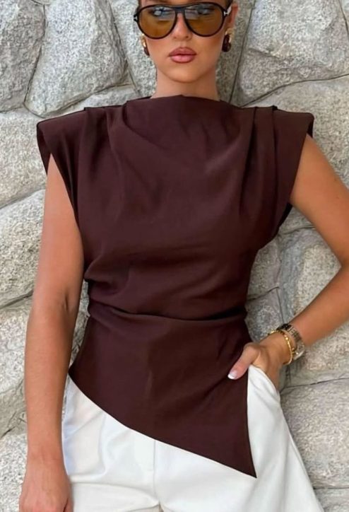 Mulher vestindo uma blusa marrom com cortes modernos, usando óculos de sol grandes, posando em frente a uma parede de pedra.