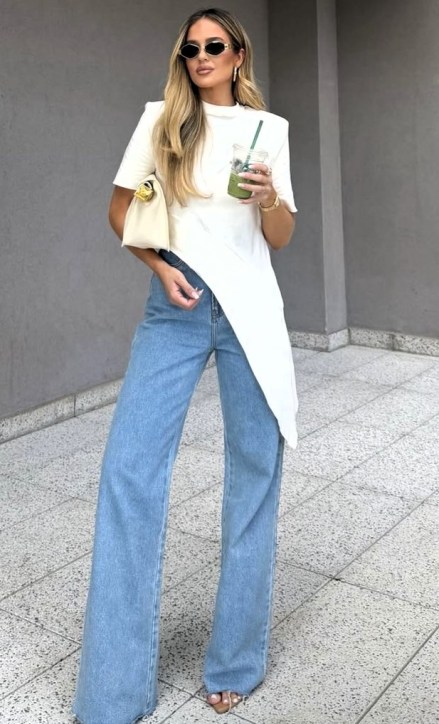 Modelo em pé com uma camiseta branca assimétrica e jeans largos, segurando uma bebida verde enquanto usa óculos escuros.