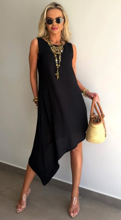 Mulher posando com um vestido preto assimétrico, usando óculos de sol e bijuterias, segurando uma bolsa de palha.