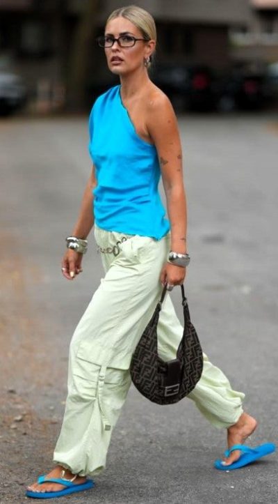 Mulher caminhando em uma rua, vestindo uma blusa azul e calças claras. Ela usa chinelos azuis e carrega uma bolsa escura.