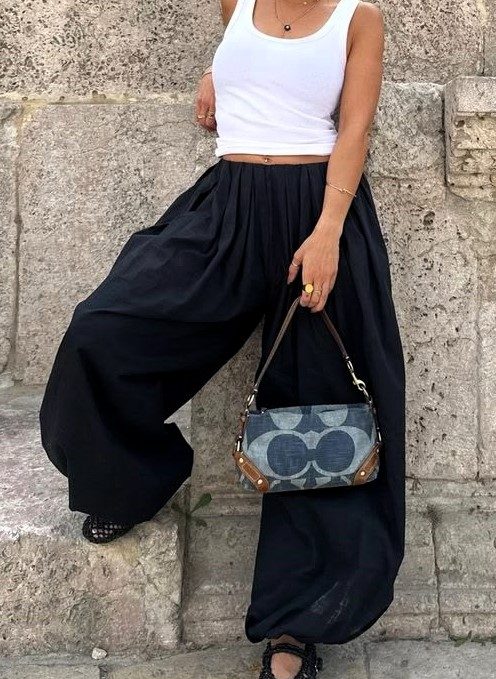 Mulher usando uma blusa branca sem mangas e calças largas pretas, segurando uma bolsa estilosa enquanto está encostada em uma parede de pedra.