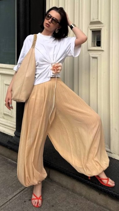 Uma mulher posando em uma rua, vestindo uma camiseta branca e calças largas de cor bege, segurando uma bolsa grande e usando sandálias vermelhas.