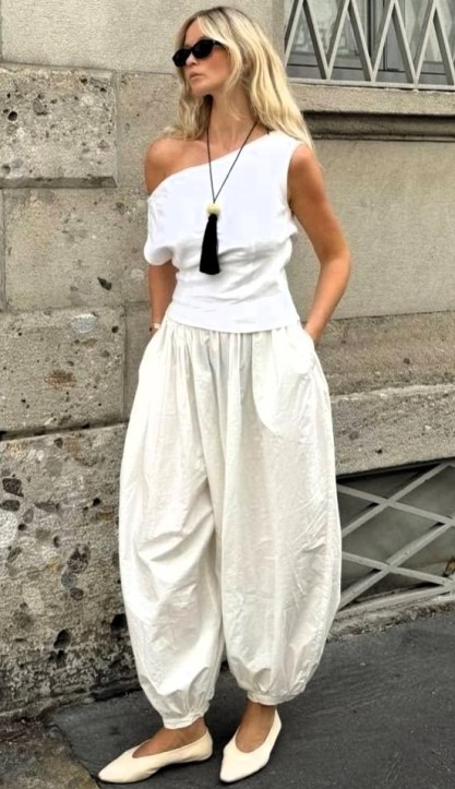 Mulher loira usando uma blusa branca de um ombro só, calças largas brancas e óculos de sol, posando em uma rua com prédios históricos ao fundo.