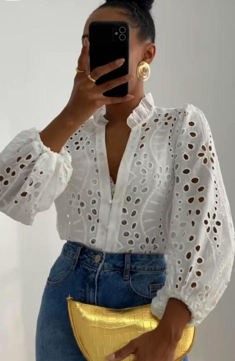 Mulher segurando um celular em um espelho, vestindo uma blusa branca com detalhes rendados e calça jeans, segurando uma bolsa amarela.
