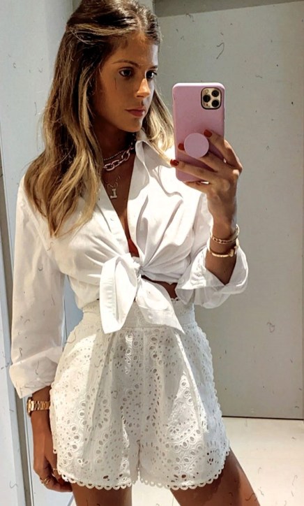 Mulher posando para o espelho com uma camisa branca amarrada na cintura e shorts brancos com detalhes de bordado.