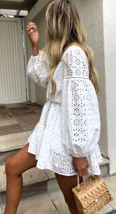 Mulher com cabelos longos e loiros, usando um vestido branco rendado, segurando uma bolsa de palha, em frente a uma entrada com piso de cerâmica.