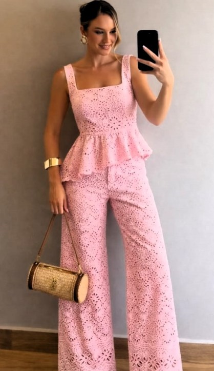 Mulher sorrindo e posando para a câmera, vestindo um conjunto rosa com detalhes em renda. Ela segura uma bolsa pequena e está com os cabelos soltos.