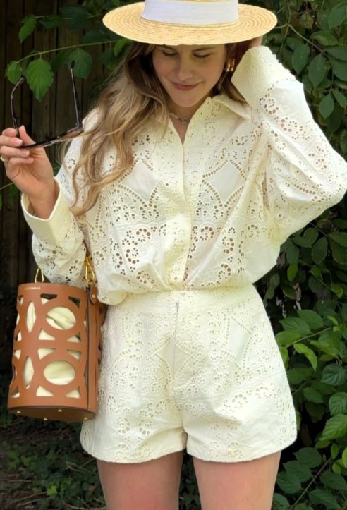 Mulher usando uma blusa e shorts brancos de renda, segurando óculos escuros e uma bolsa de design moderno, com um chapéu de palha, em um ambiente natural com folhagens ao fundo.