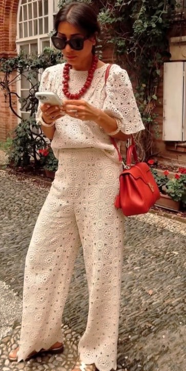 Mulher usando um conjunto de roupas brancas de renda, segurando um celular e com óculos escuros, ao fundo uma parede de tijolos e flores.