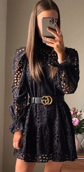 Mulher tirando uma selfie com um vestido preto de renda, mostrando detalhes de cortes circulares e um cinto preto com fivela dourada.