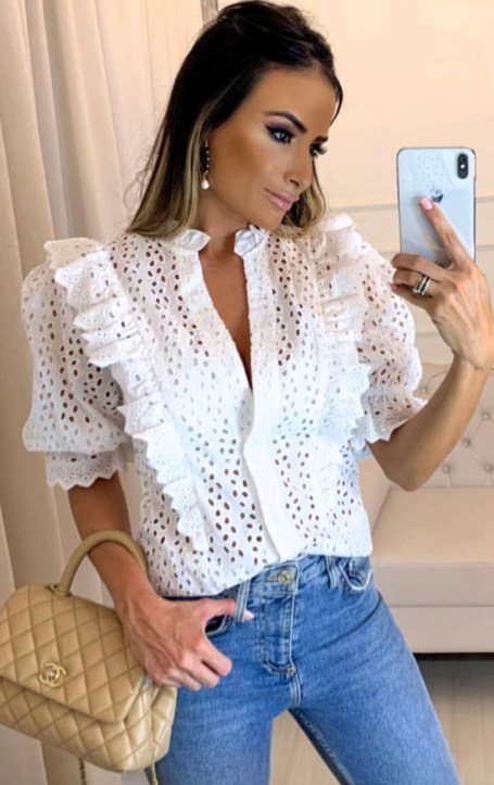 Mulher tirando selfie no espelho, vestindo uma blusa branca com babados e jeans. Ela segura uma bolsa de designer.