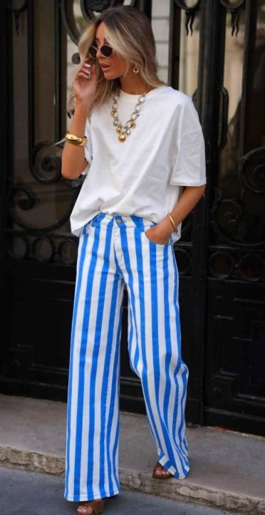Pessoa com cabelo longo e loiro, usando uma camiseta branca e calças listradas em azul e branco. Ela está de pé, com uma mão no cabelo e a outra na bolsa, usando óculos escuros e acessórios dourados. Ao fundo, uma porta com um design decorativo.