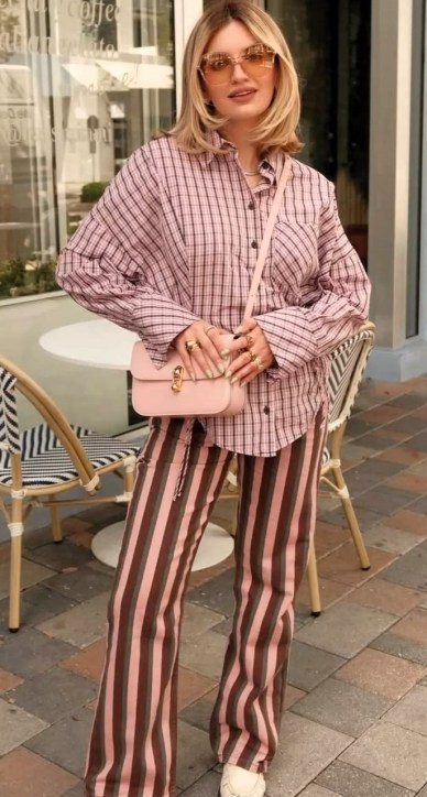 Mulher posando em um ambiente externo, usando uma camisa de xadrez rosa e calças listradas em tons de rosa. Ela segura uma bolsa rosa e usa óculos de sol. Há mesas e cadeiras ao fundo.