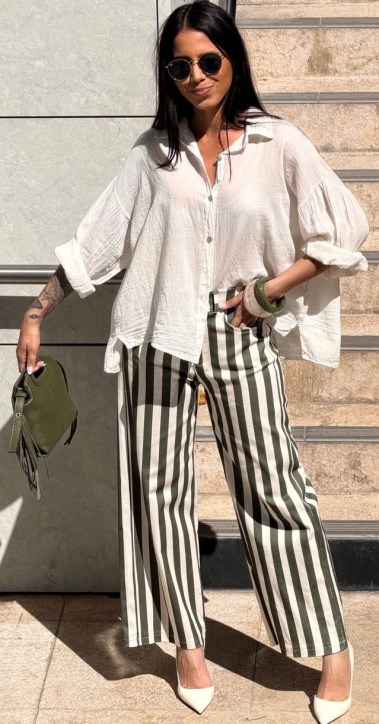 Mulher com camisa branca de manga larga e calças listradas em preto e verde, usando óculos escuros e segurando uma bolsa verde.