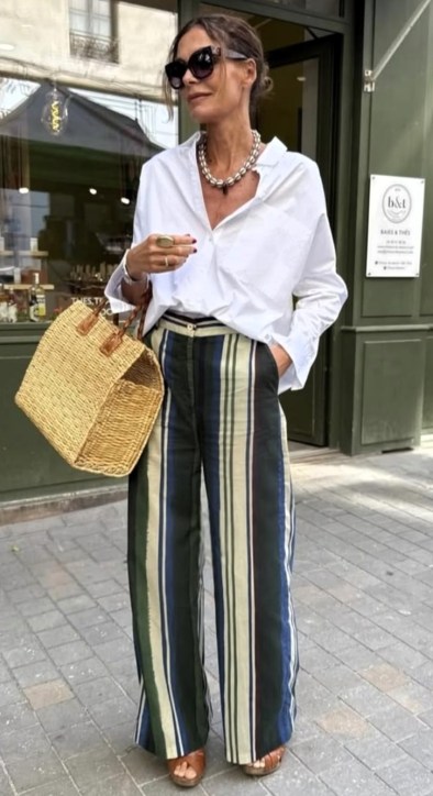 Uma mulher stylish usando uma camisa branca oversized e calças listradas largas, segurando uma bolsa de palha e usando óculos escuros.