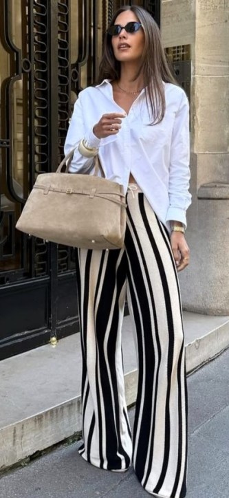 Mulher vestindo uma camisa branca e calças listradas em preto e branco, segurando uma bolsa bege. Ela usa óculos escuros e está em um ambiente urbano.