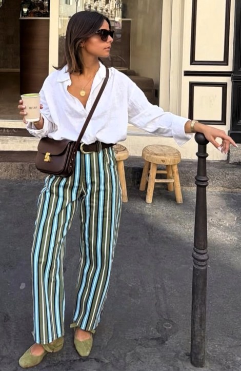 Mulher em pé usando uma camisa branca e calças listradas em verde, azul e branco, segurando um copo de café. Ela usa óculos escuros e uma bolsa marrom, com um fundo de rua e bancos de madeira.