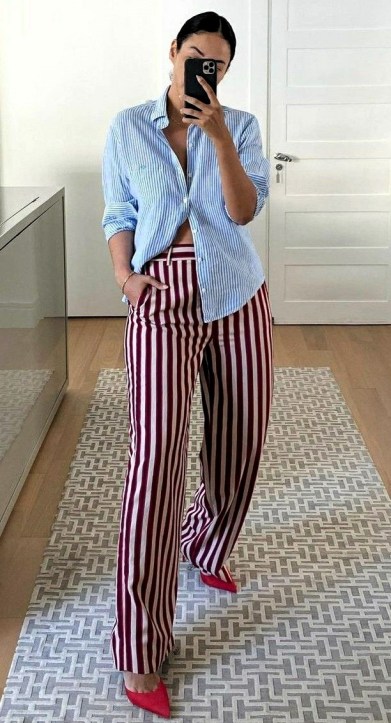 Pessoa em pé, refletindo em um espelho, vestindo uma camisa azul listrada e calças largas listradas em vermelho e branco, com sapatos de salto alto rosa.