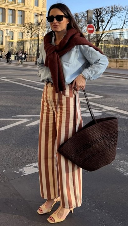 Mulher em pé na rua usando uma camisa jeans azul e calças listradas em tons de marrom e bege, segurando uma bolsa grande e com um cachecol vermelho ao redor do pescoço.
