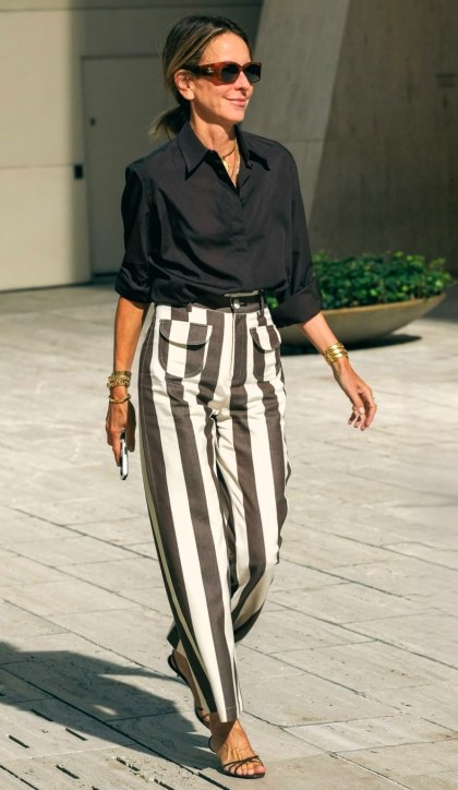 Mulher elegante caminhando em um ambiente urbano, vestindo uma camisa preta e calças listradas em preto e branco.