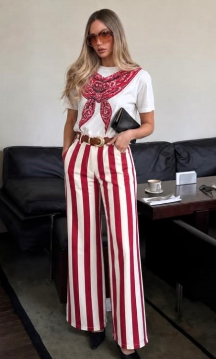 Mulher com camisa branca e lenço estampado, usando calças largas listradas em vermelho e branco, segurando uma bolsa preta, em um ambiente moderno.