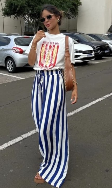 Uma mulher usando uma camiseta branca com estampa colorida e uma calça listrada azul e branca, andando em um estacionamento.