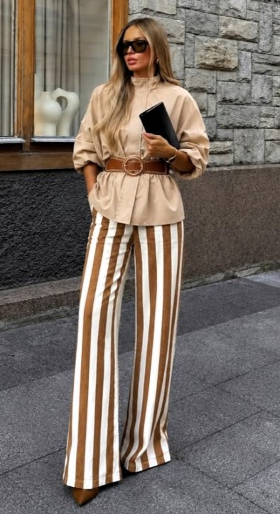 Mulher com blusa clara, cinto marrom e calças listradas em tons de marrom e branco, usando óculos escuros e segurando uma bolsa preta, posando em ambiente urbano.