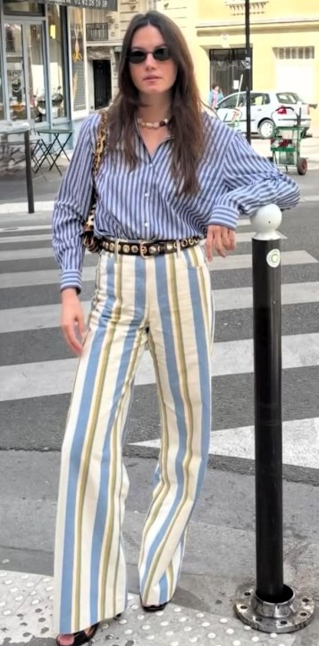 Jovem usando camisa listrada azul e branca e calças listradas em tons de azul e bege, posando na calçada ao lado de um poste.