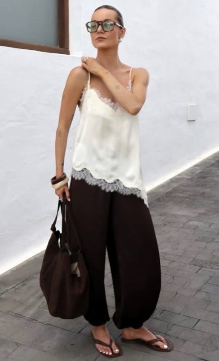 Mulher vestindo blusa de alça branca com renda e calças largas marrons, segurando uma bolsa marrom e usando óculos escuros, em uma parede clara.