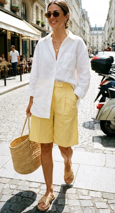 Mulher sorridente, vestindo uma camisa branca de linho e bermuda amarela, segurando uma bolsa de palha, caminhando por uma rua de paralelepípedos com mesas ao ar livre ao fundo.