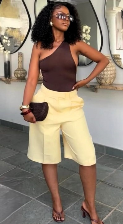 Mulher com cabelo cacheado usa um body marrom e bermudas amarelas, segurando uma bolsa escura em um ambiente elegante.