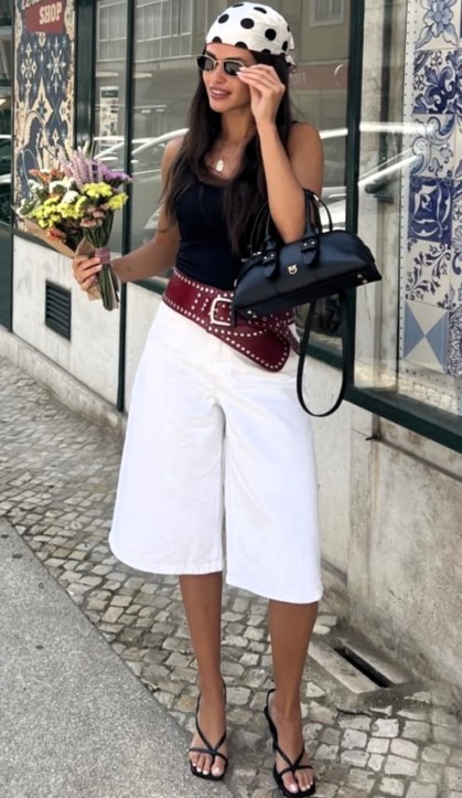 Mulher usando uma blusa preta e calças brancas largas, segurando flores e uma bolsa preta, com um lenço de bolinhas na cabeça.