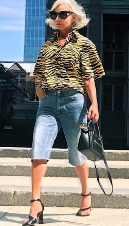 Mulher com cabelo platinado usando uma camisa de estampa de tigre, shorts jeans e sandálias de salto alto, caminhando em um ambiente urbano.