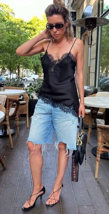 Mulher com blusa preta de alças finas e detalhes em renda, usando shorts jeans rasgado e sandálias de tiras. Ela está em um ambiente ao ar livre, próximo a mesas de restaurante e árvores.