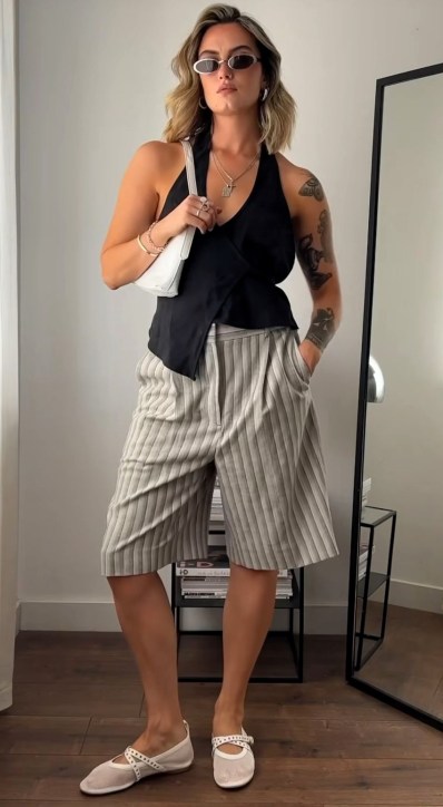 Mulher de pé em um ambiente interno, vestindo uma blusa preta e bermuda listrada. Ela usa óculos de sol e acessórios, e segura uma bolsa branca. No fundo, há um espelho e um chão de madeira.