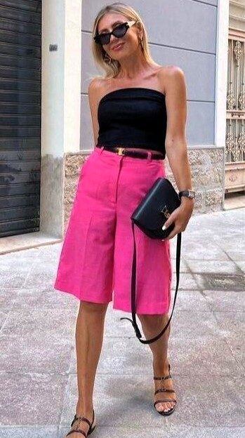 Mulher usando blusa preta sem alças e bermuda rosa, segurando uma bolsa preta, caminhando em uma calçada.