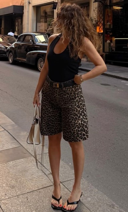 Mulher com cabelo cacheado, vestindo uma camiseta preta e bermuda de estampa de onça, segurando uma bolsa branca, em uma rua urbana.