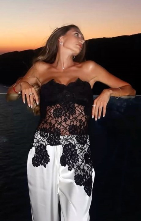 Mulher com blusa de rendas pretas e calças brancas, posando em um local ao ar livre com o mar e o pôr do sol ao fundo.