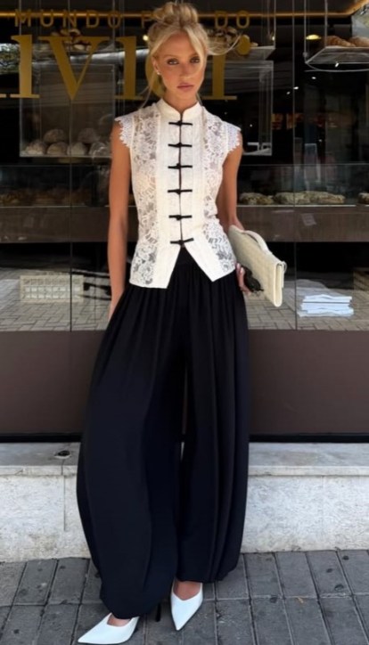Modelo feminina usando uma blusa de renda branca e calças largas pretas na frente de uma padaria.