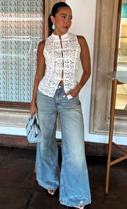 Mulher posando em frente a uma parede de vidro. Ela usa uma blusa branca de renda e jeans boca de sino. Acessórios incluem brincos grandes e uma bolsa azul. O ambiente é bem iluminado.