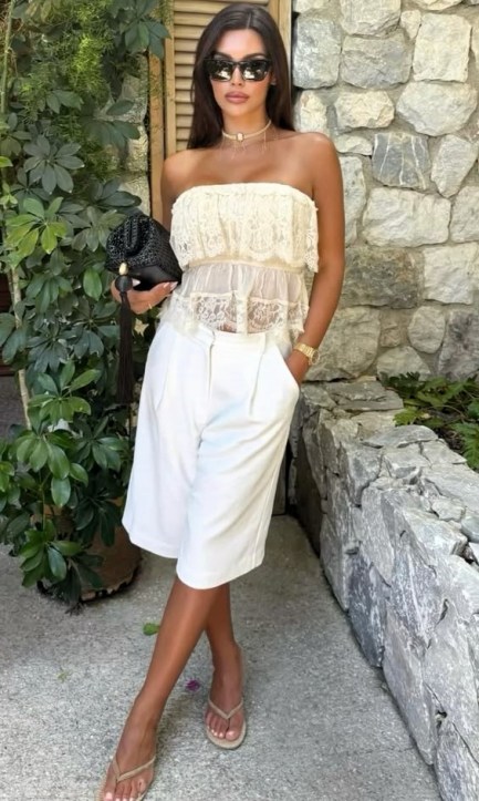 Mulher em um ambiente externo, usando uma blusa strapless de renda branca, calças curtas brancas e sandálias, segurando uma bolsa preta, com plantas verdes e uma parede de pedras ao fundo.