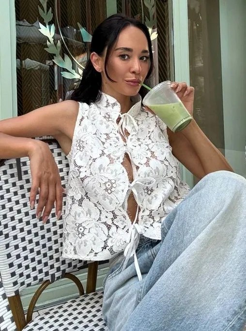 Mulher com blusa de renda branca sentada em uma cadeira, segurando um copo de bebida verde.