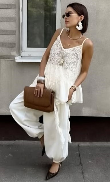Mulher vestindo blusa branca de renda e calças brancas, segurando uma bolsa marrom e usando óculos de sol.