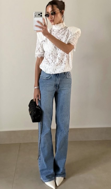 Mulher posando em frente ao espelho, vestindo uma blusa de renda branca e jeans claros. Ela segura uma bolsa preta e usa óculos escuros.