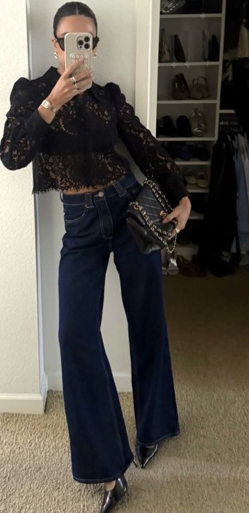 Mulher posando com blusa de renda preta e calças jeans largas, segurando uma bolsa e tirando uma selfie em frente a um espelho.