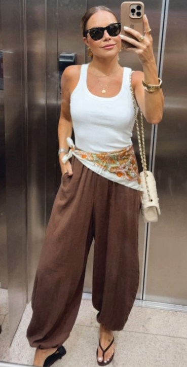Mulher na frente do espelho segurando o celular, usando uma blusa branca, calças largas marrons e sandálias. Ela também usa óculos escuros e uma bolsa clara com detalhes.
