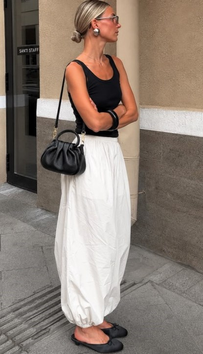 Mulher de cabelo preso em um coque, usando blusa preta e saia branca longa, segurando uma bolsa preta, posando em uma calçada.